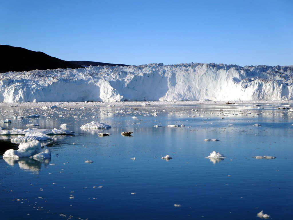 The white ice wall of the Eqip Sermia glacier across calm blue water with small floating ice pieces under a clear sky