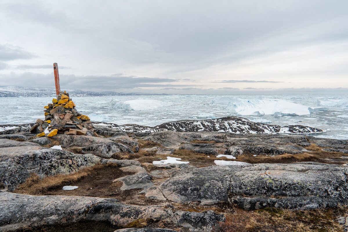 A stone cairn with a red pole on bare rock, with sea ice and icebergs stretching to the horizon