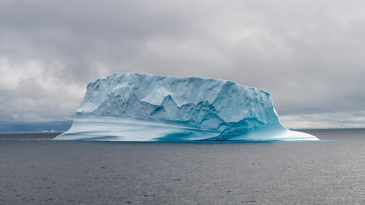 A large flat-topped iceberg with blue-white ice floating in calm grey water of Disko Bay under heavy clouds
