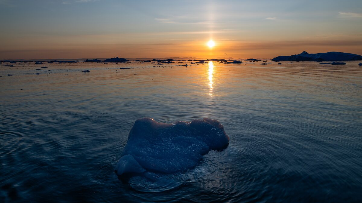 The midnight sun low over Disko Bay, with a small iceberg floating in the dark blue water