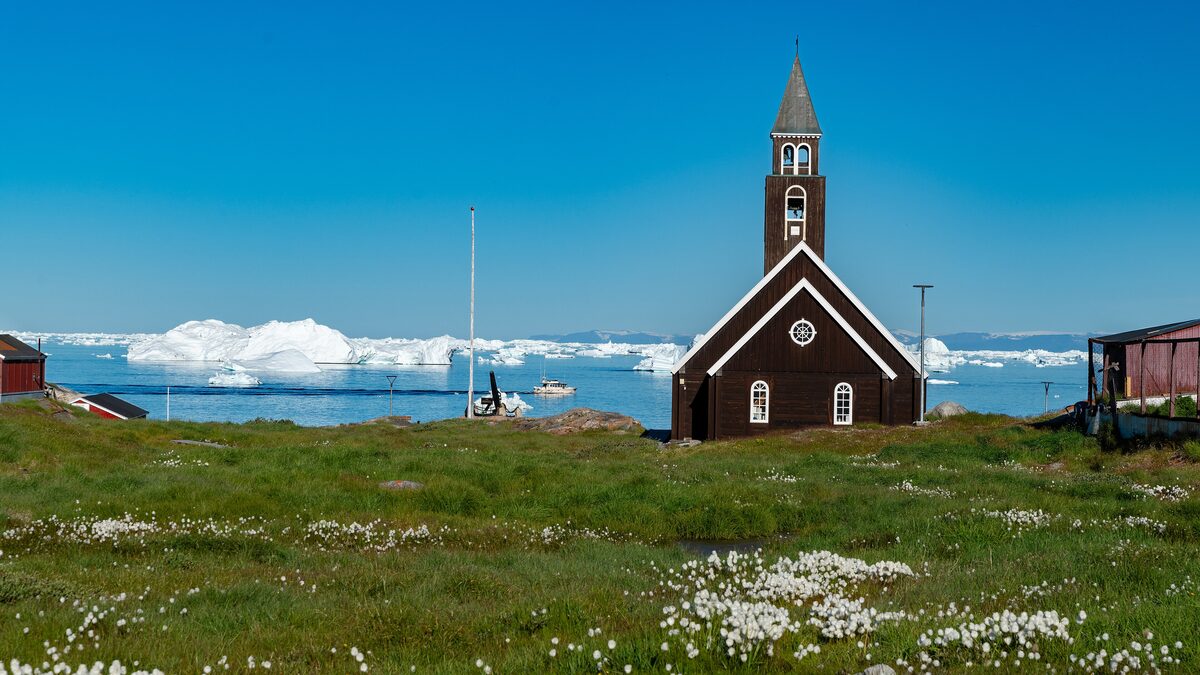 Zion Church, a dark red wooden church on green grass at the edge of Ilulissat, with icebergs on the water behind it