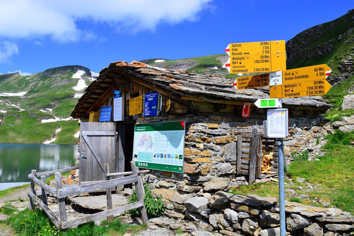 A stone-and-timber hut with trail signposts beside the Bachalpsee, with the lake and mountains behind