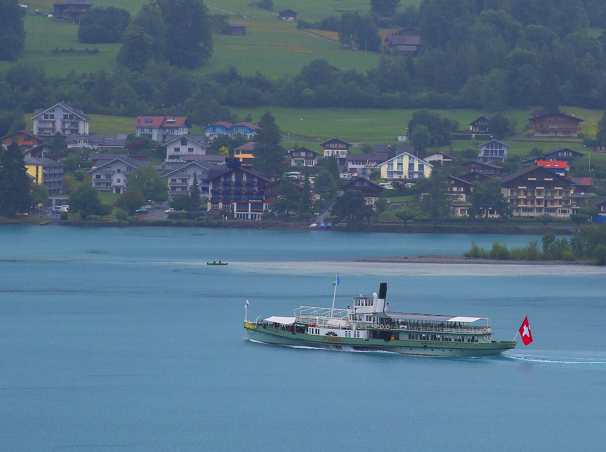 The green paddle steamer Lötschberg crossing Lake Brienz with a Swiss flag at the stern, and lakeside houses on the shore behind