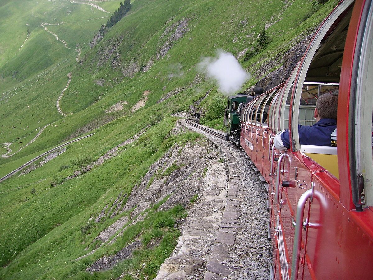 A red steam train climbing a grassy hillside on the Brienz Rothorn Railway