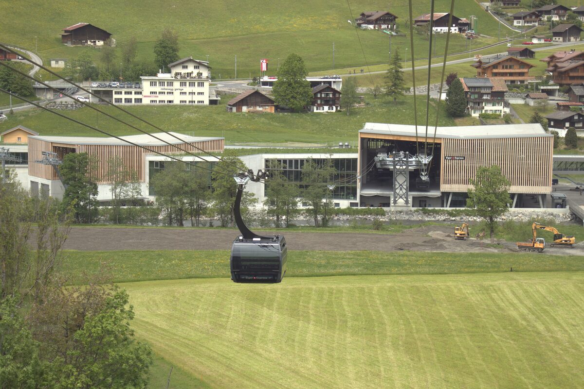The Eiger Express gondola gliding in over green meadows toward the Grindelwald Terminal building, with chalets on the hill behind