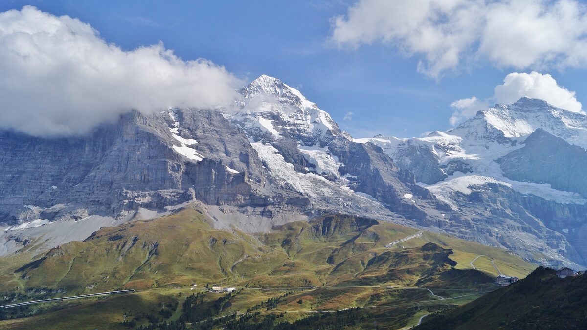 The Eiger and Mönch rising above green alpine pastures and a grassy ridge, with clouds over the summits