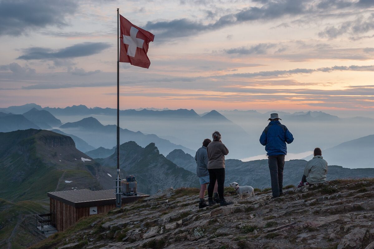 Hikers by the Swiss flag on the Faulhorn in the evening light, with ridges fading into haze