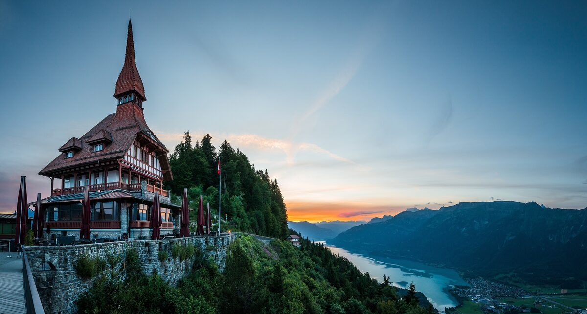 The Harder Kulm restaurant on its stone-walled terrace at dusk, with an orange glow on the horizon above a lake and the valley below