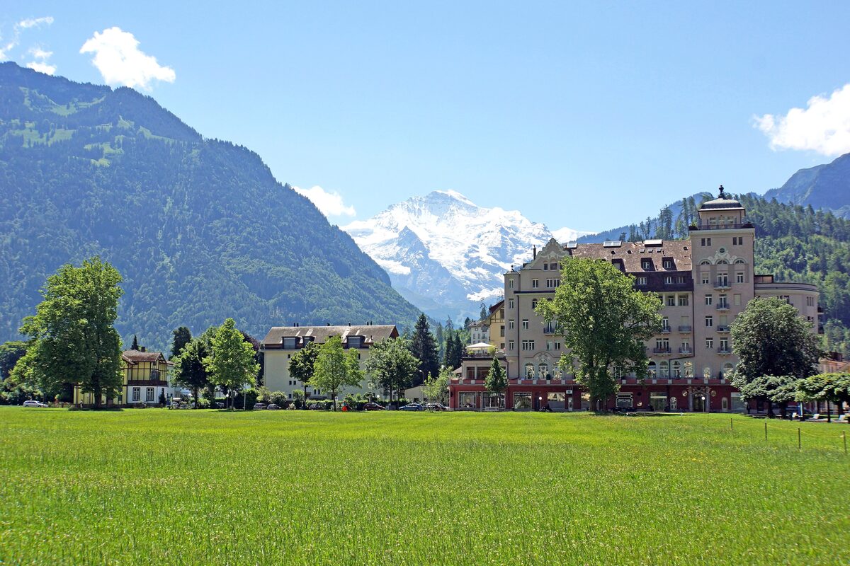 The green Höhematte meadow in Interlaken with a grand hotel at its edge and the snowy Jungfrau rising beyond the forested slopes