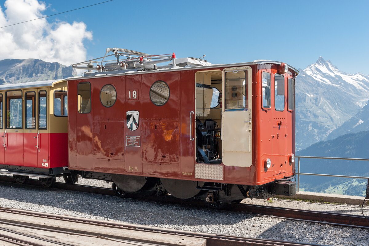 A red vintage electric rack locomotive, number 18, with cars behind it at the Schynige Platte top station, a snowy peak rising beyond