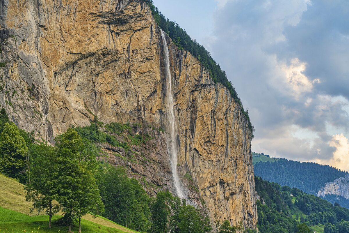 The Staubbach Falls dropping down a sheer cliff above green meadows and pine trees in summer