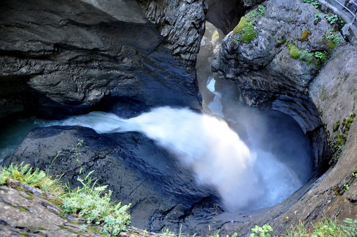 Fast meltwater pouring out of a dark rock gorge at Trümmelbach Falls, with moss and grass on the wet stone
