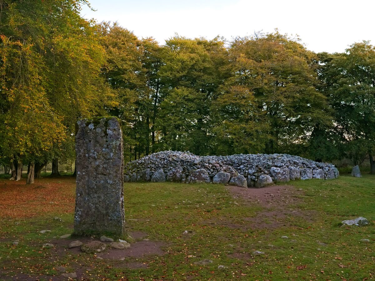 A standing stone and a round Bronze Age burial cairn of piled stones among autumn trees at Clava