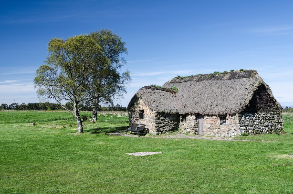 Leanach Cottage, a small thatched stone cottage on the open moor of the Culloden battlefield, with two trees beside it