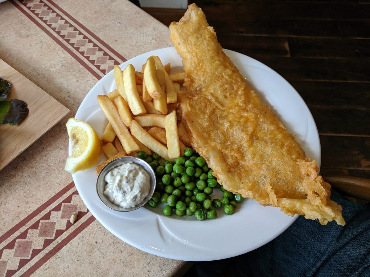 A long piece of battered haddock with chips, peas, lemon and tartare sauce on a plate