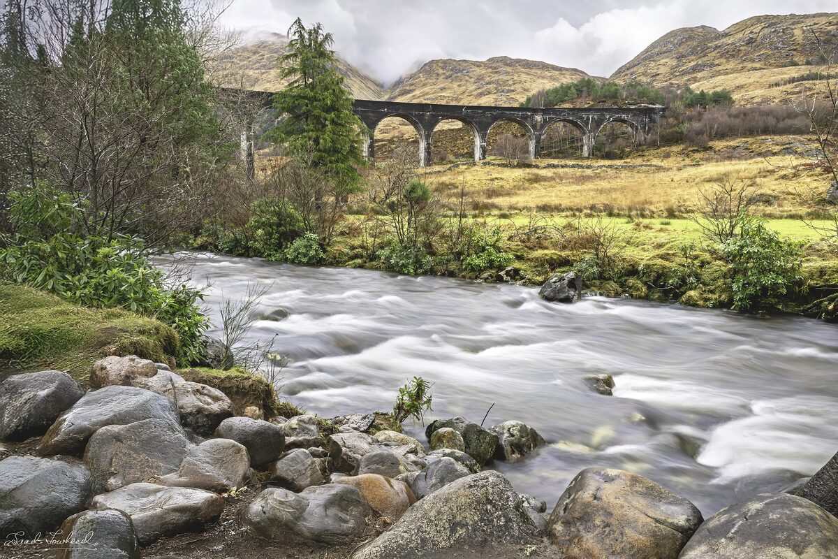 The curving arches of the Glenfinnan Viaduct across a green glen, with a rushing river in front