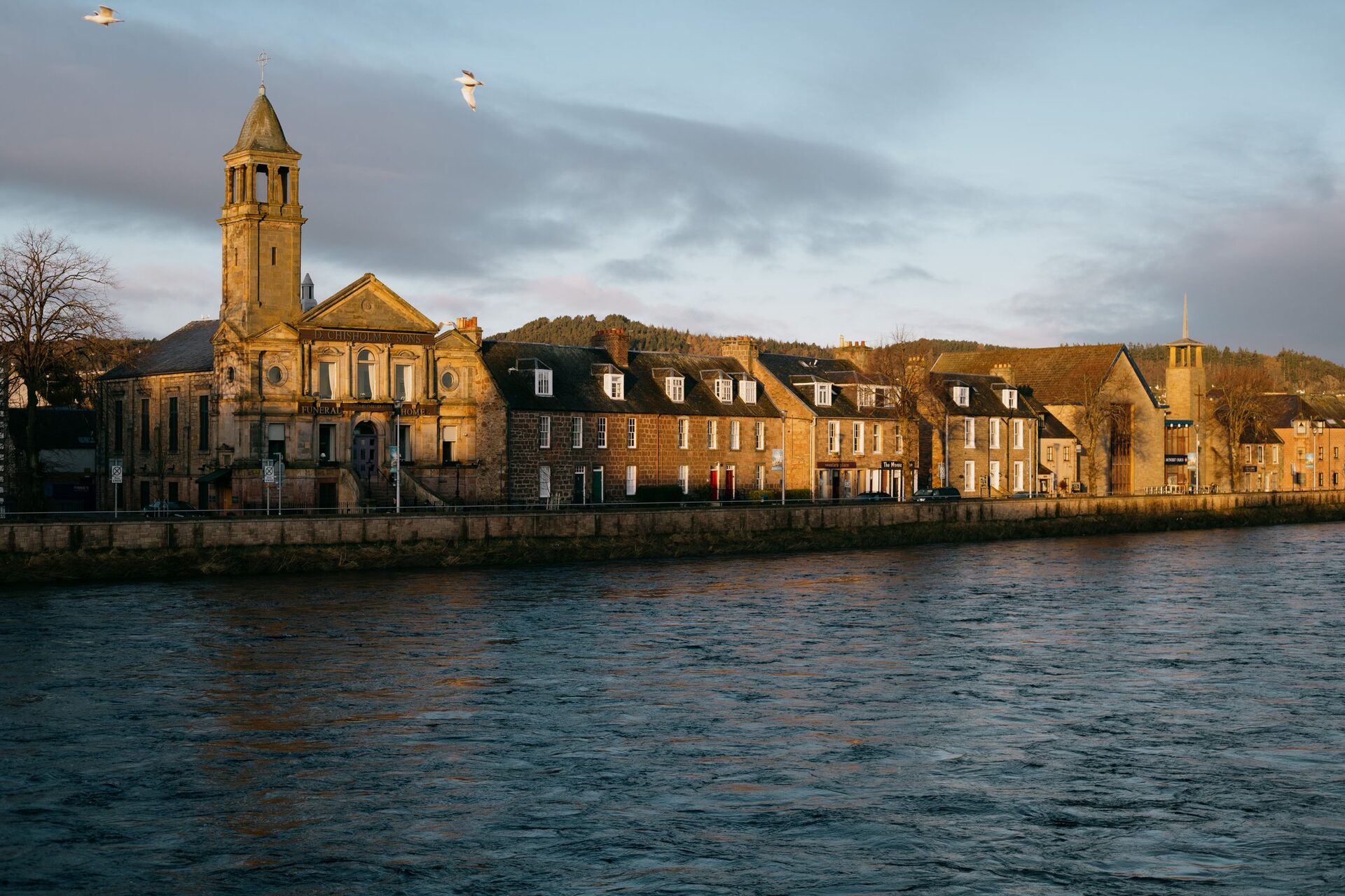 The River Ness in Inverness at sunrise, with a church tower and a row of old stone houses along the far bank