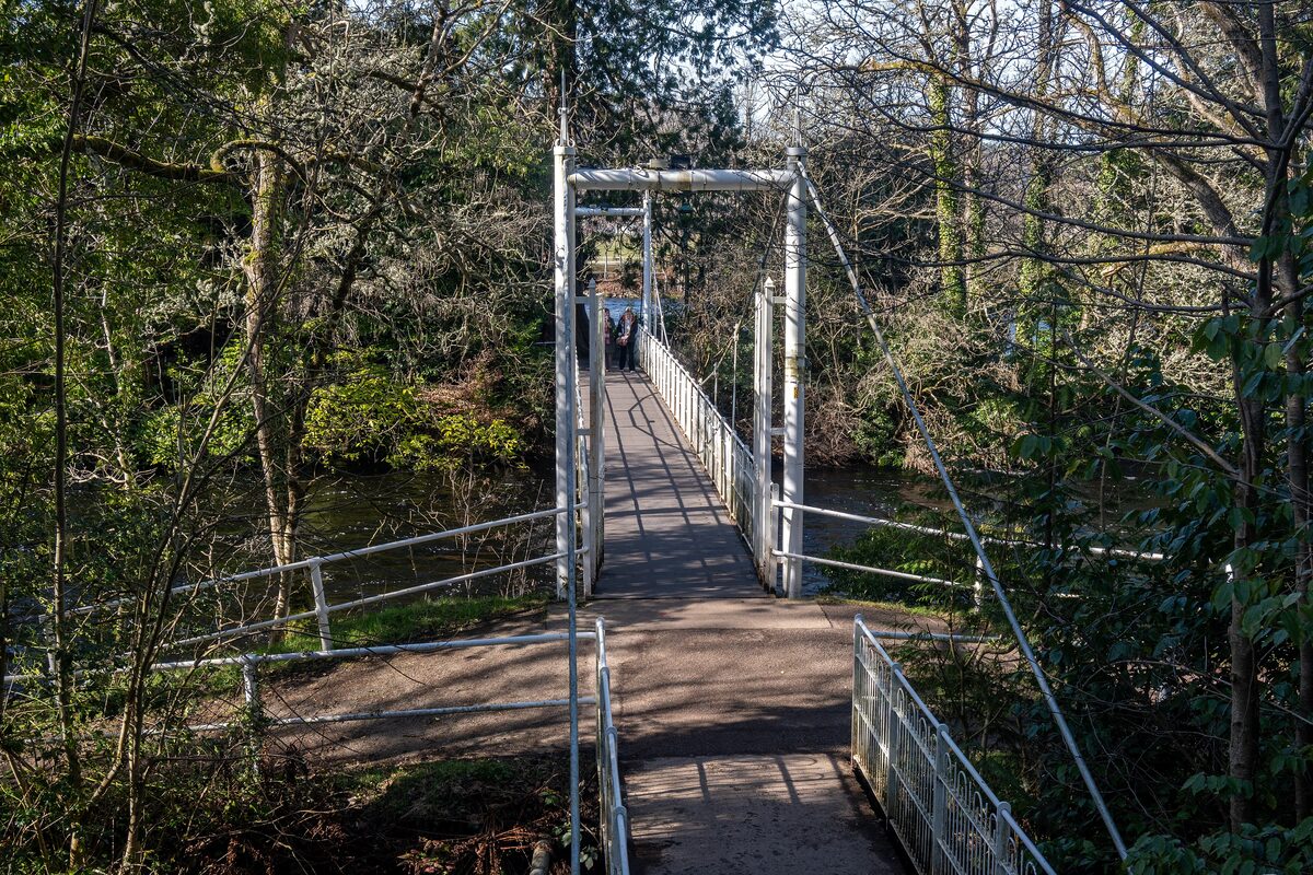 A white suspension footbridge over the River Ness among the trees of the Ness Islands