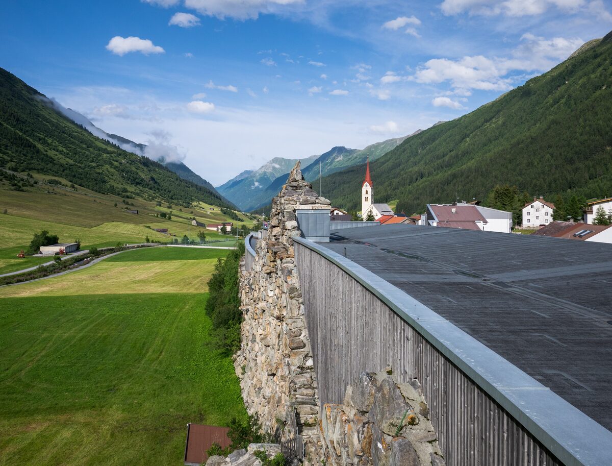 The long timber-clad roof of the Alpinarium built into its stone protection wall, with green meadows, the Galtür church and a valley behind (summer)