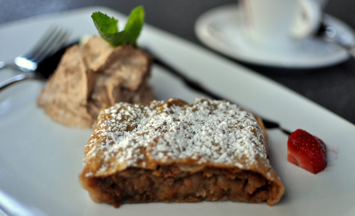 A slice of apple strudel dusted with icing sugar, with a scoop of cream, a strawberry and a cup of coffee