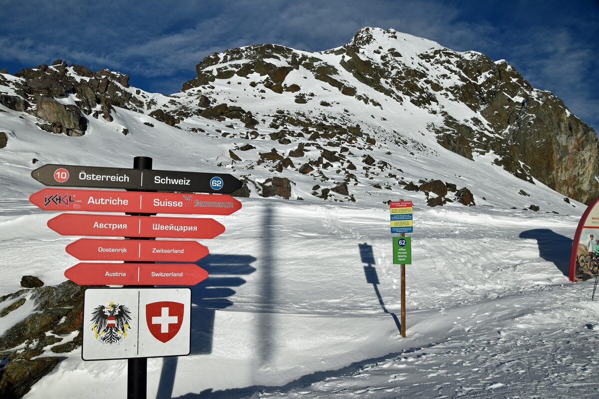 A signpost pointing to Austria and Switzerland (Samnaun) with the Austrian and Swiss coats of arms, on snow below a rocky summit