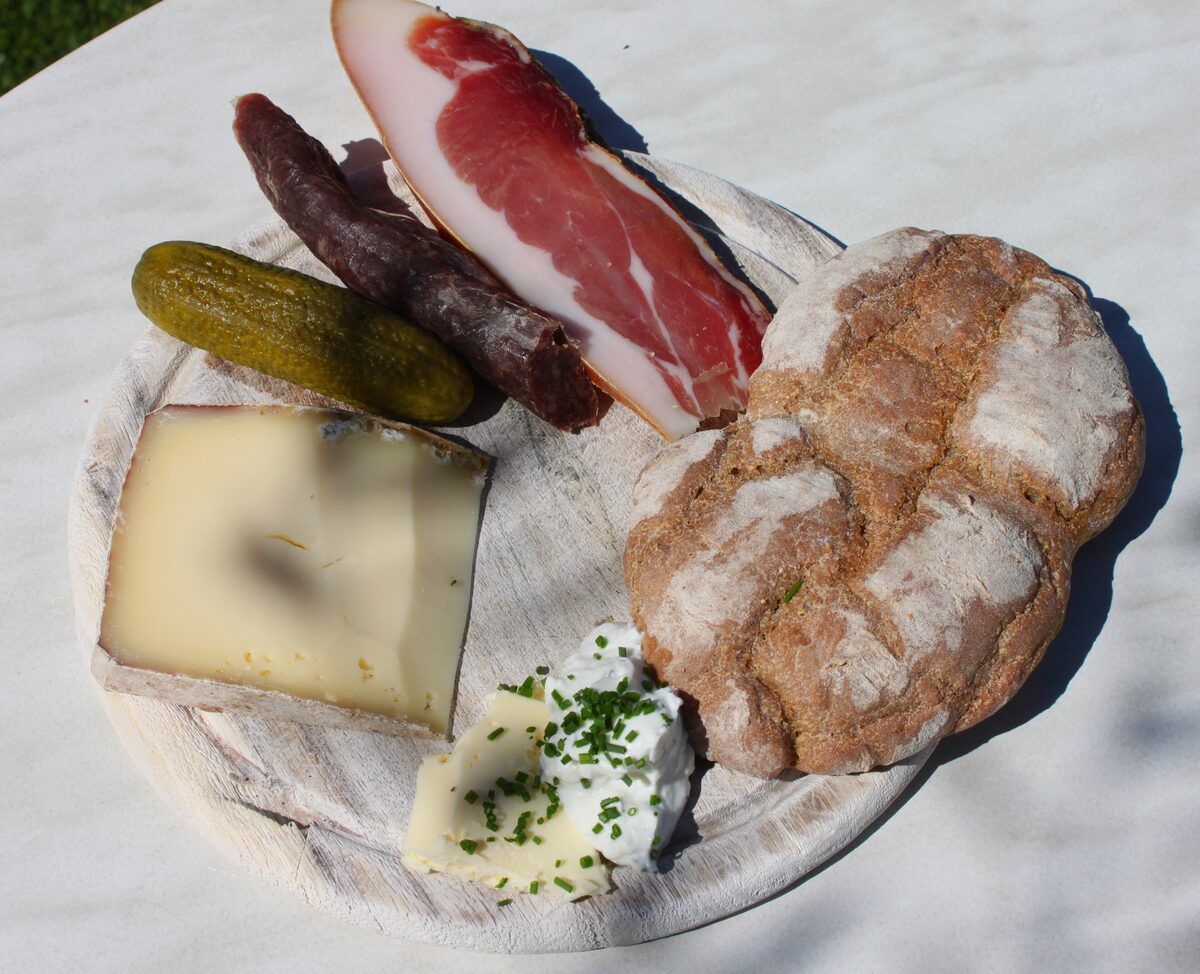 A wooden board with cured ham, a smoked sausage, a gherkin, a wedge of cheese, herb cream and a round of dark bread