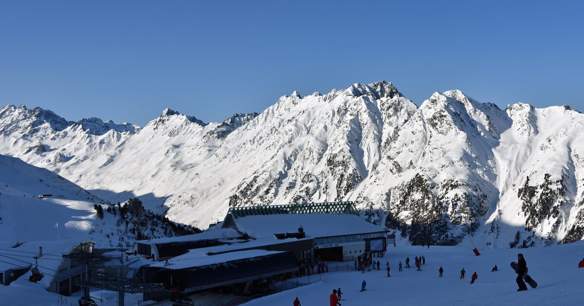 The Idalp mountain station in the Silvretta Arena with skiers and a snowboarder under snowy peaks on a clear winter day