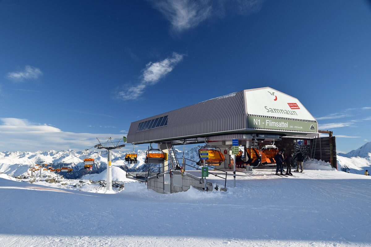 The Flimsattel top station with a Samnaun sign on its roof, skiers and lifts on a sunny winter day