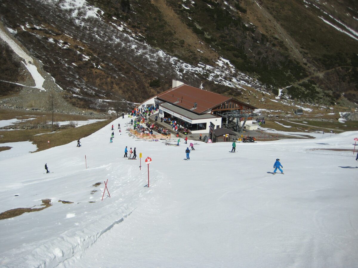 A mountain restaurant with a busy terrace above a thin, patchy spring piste with a few skiers and snowboarders