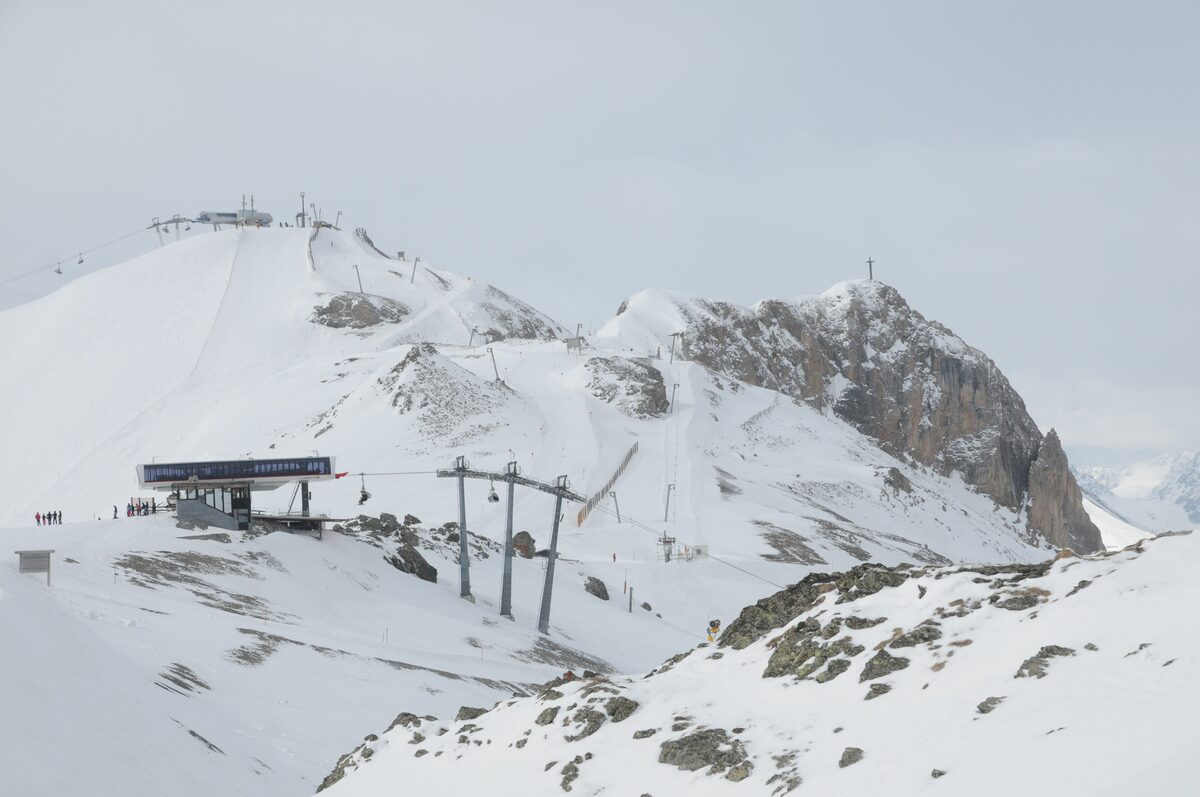 A lift station on a snowy ridge below a rocky summit with a cross, under an overcast sky