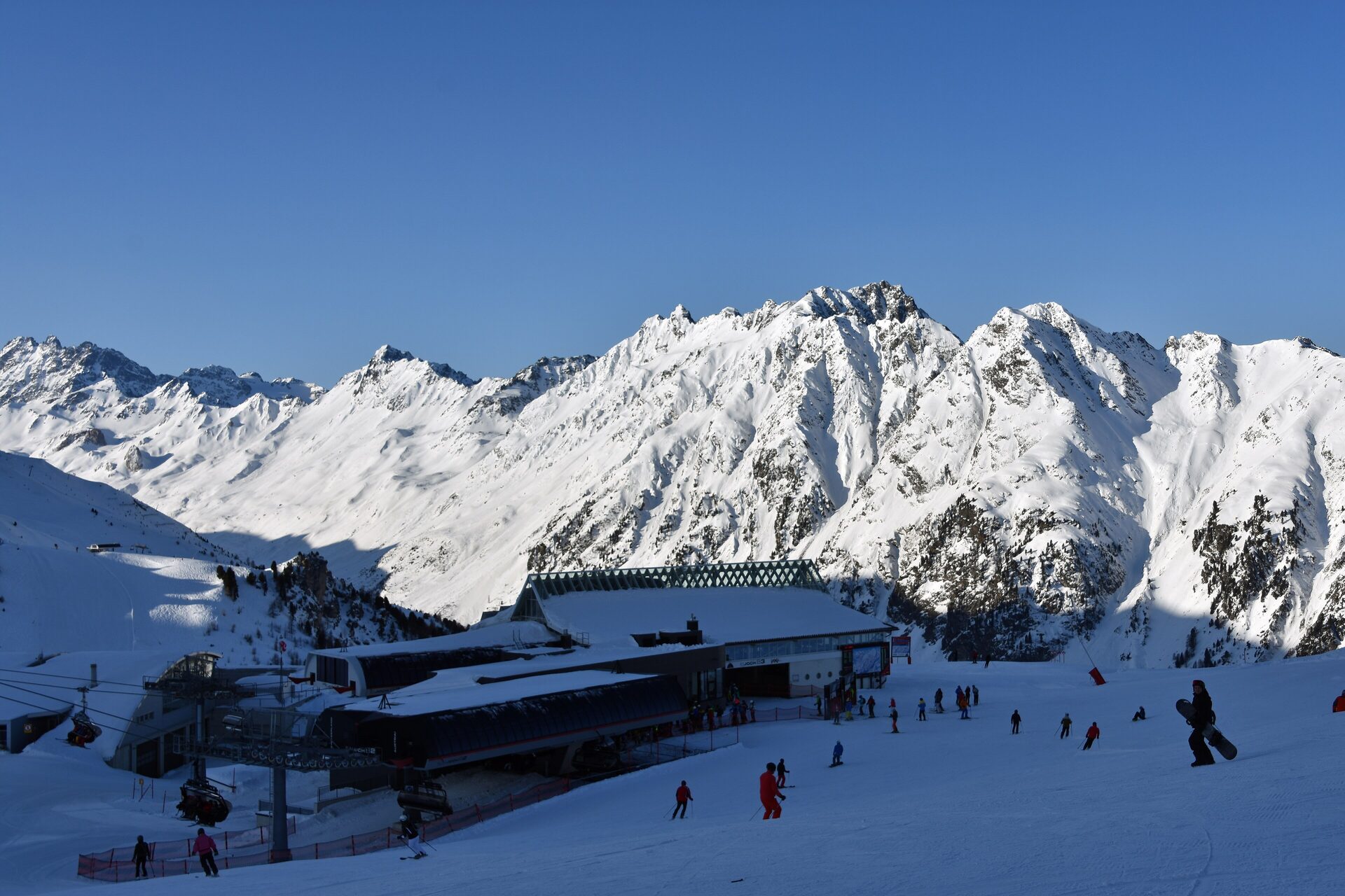 The Idalp mountain station in the Silvretta Arena with skiers and a snowboarder under snowy peaks on a clear winter day