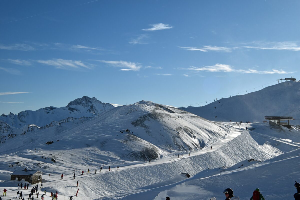 A wide snowy piste with a few skiers below mountain ridges under a blue sky