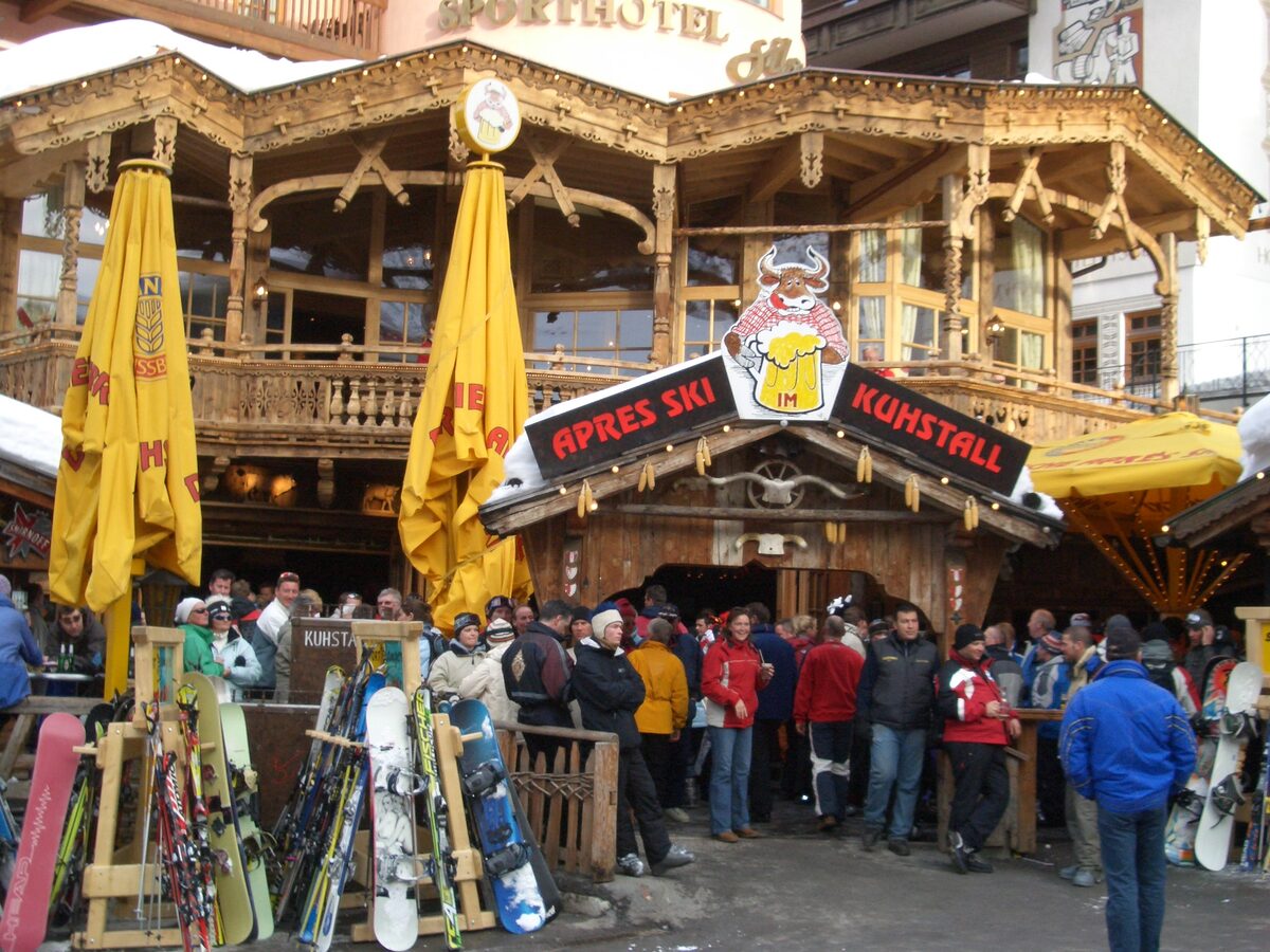 A crowd of skiers and snowboarders outside the timber Kuhstall après-ski bar in Ischgl, with skis in racks and yellow parasols
