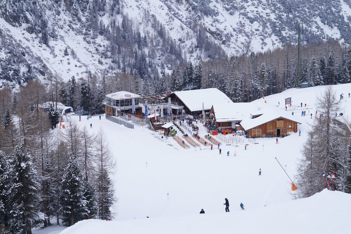 Mountain huts with a terrace beside a snowy piste, with skiers below and pine trees and a steep slope behind