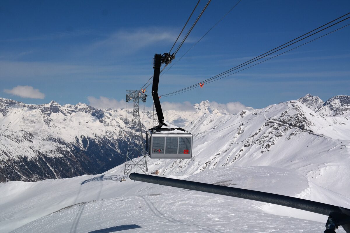 A gondola cabin on the Piz Val Gronda lift with snowy peaks and a blue sky behind