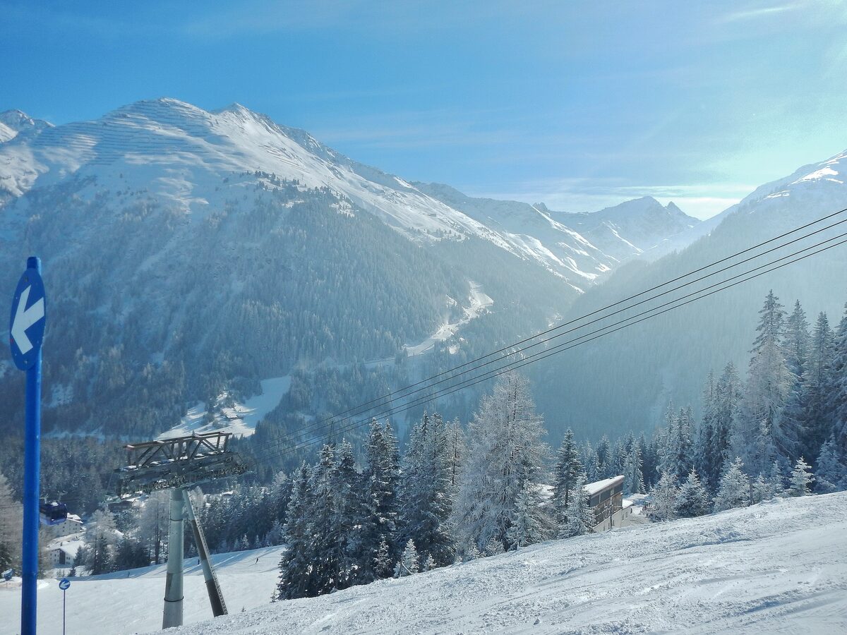A snowy piste with frosted pines and a lift pylon above the Arlberg valley, with sunlit mountains behind