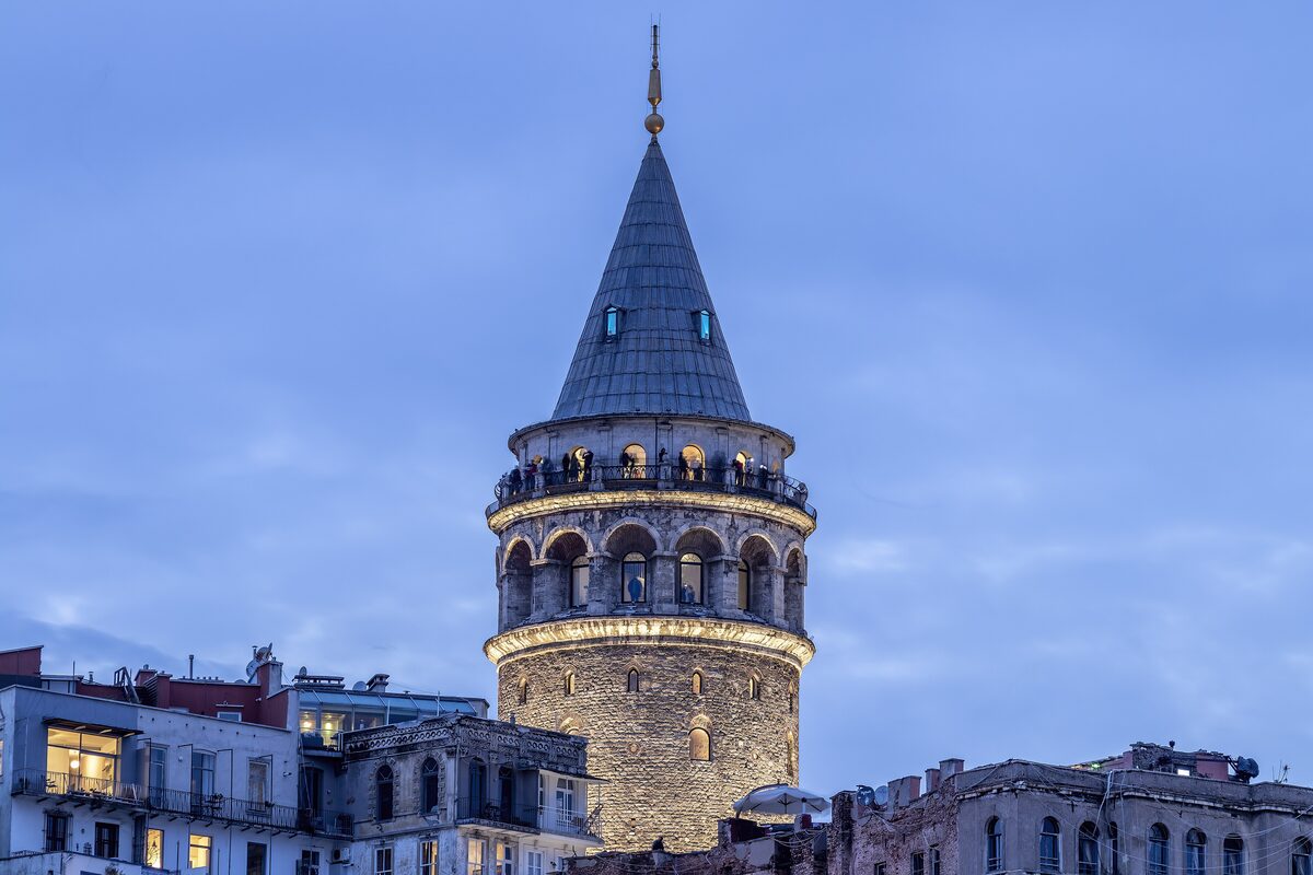 The stone Galata Tower with its conical roof and lit gallery at dusk, above the rooftops of Galata