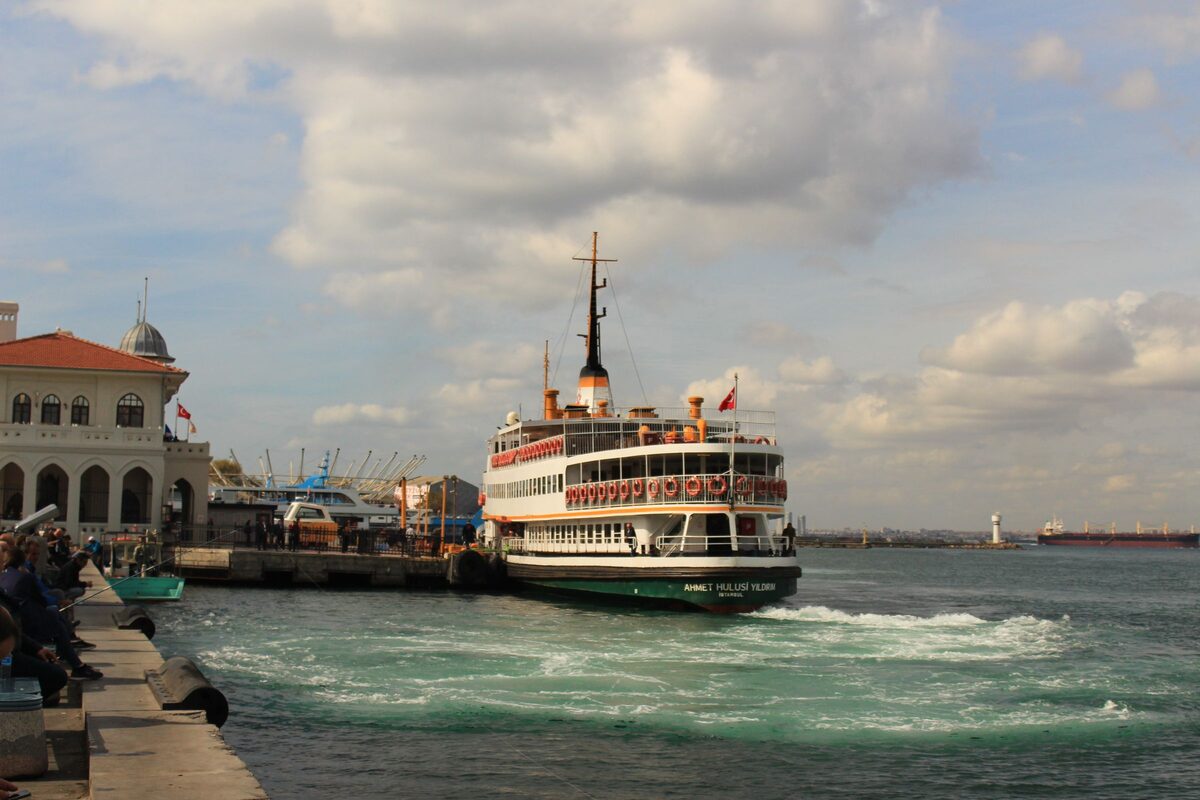 A white city ferry leaving the Kadıköy pier under a cloudy sky
