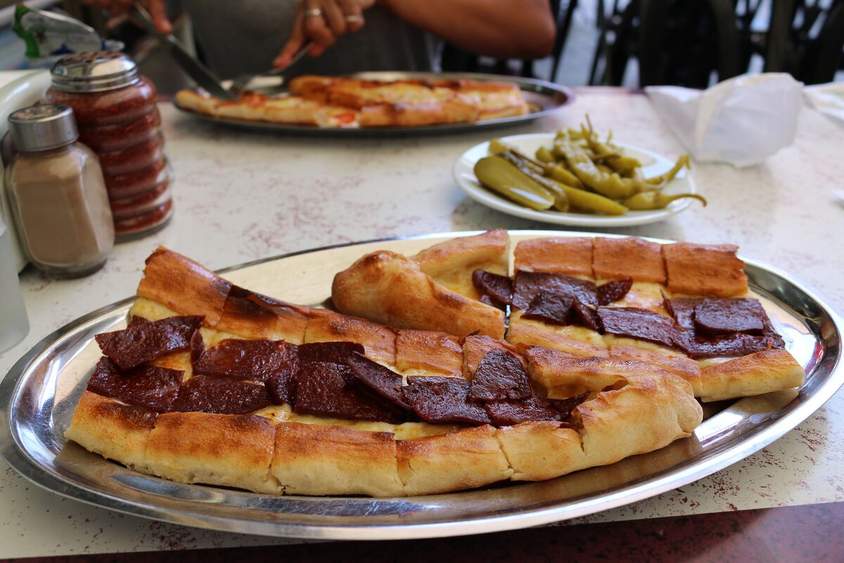 A boat-shaped pide topped with slices of sucuk sausage, on a metal platter with pickled peppers