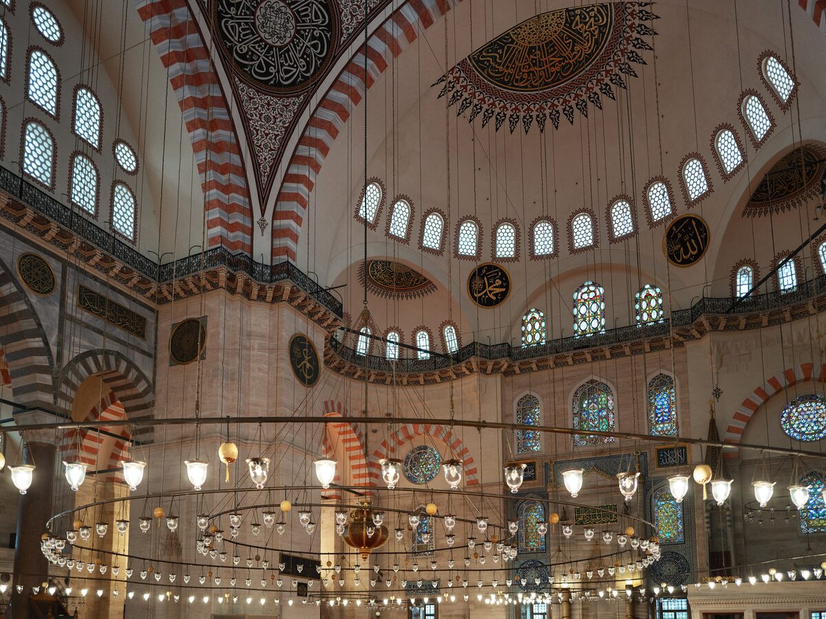 Inside the Süleymaniye Mosque: red and white striped arches, rows of windows, the painted dome and a ring of hanging lamps