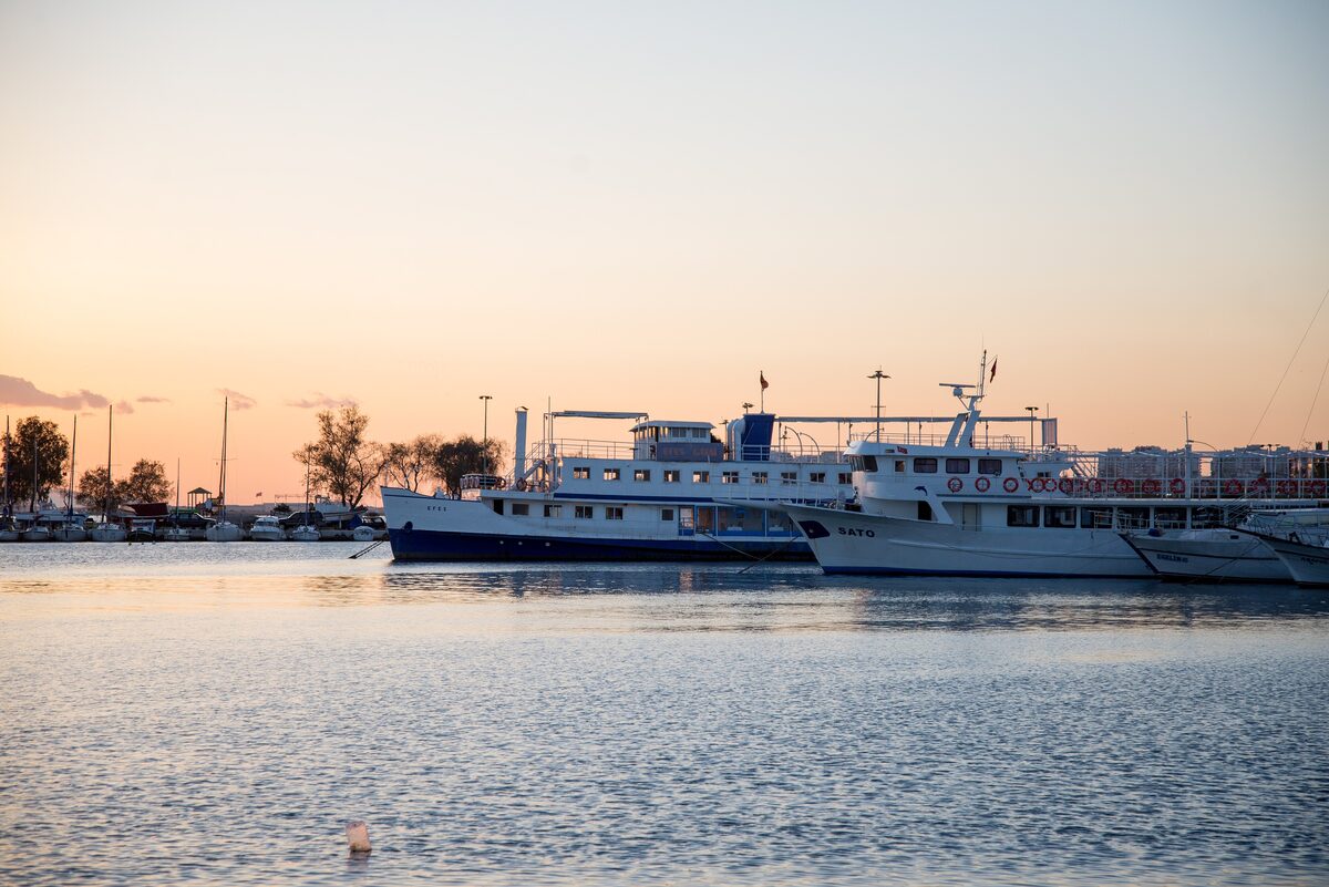 Ferries and fishing boats moored in the harbor at Bostanlı at sunset, with the sky turning orange behind them