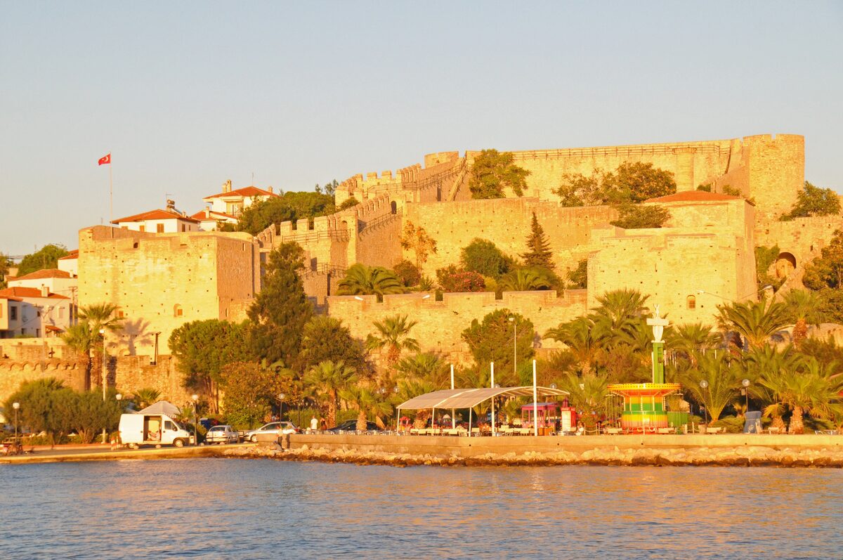 The stone walls and towers of Çeşme Castle glowing in late afternoon sun above the palm-lined waterfront