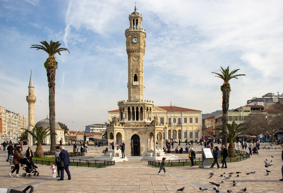 The İzmir Clock Tower in Konak Square, a slender carved stone tower on a round fountain base, between palm trees with the minaret of Konak Mosque on the left