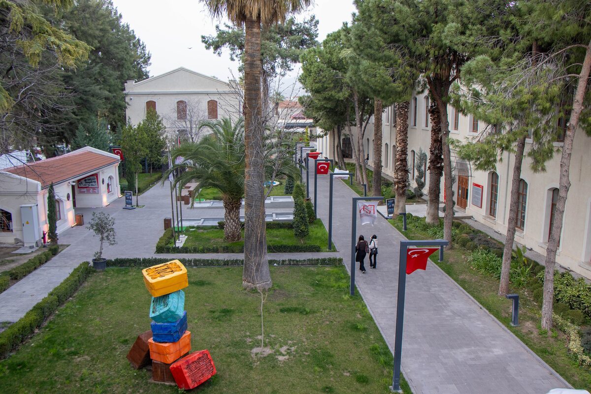 Palm trees, lawns and a sculpture of stacked colorful crates in the garden of the İzmir Culture and Art Factory, with the old whitewashed factory buildings around it