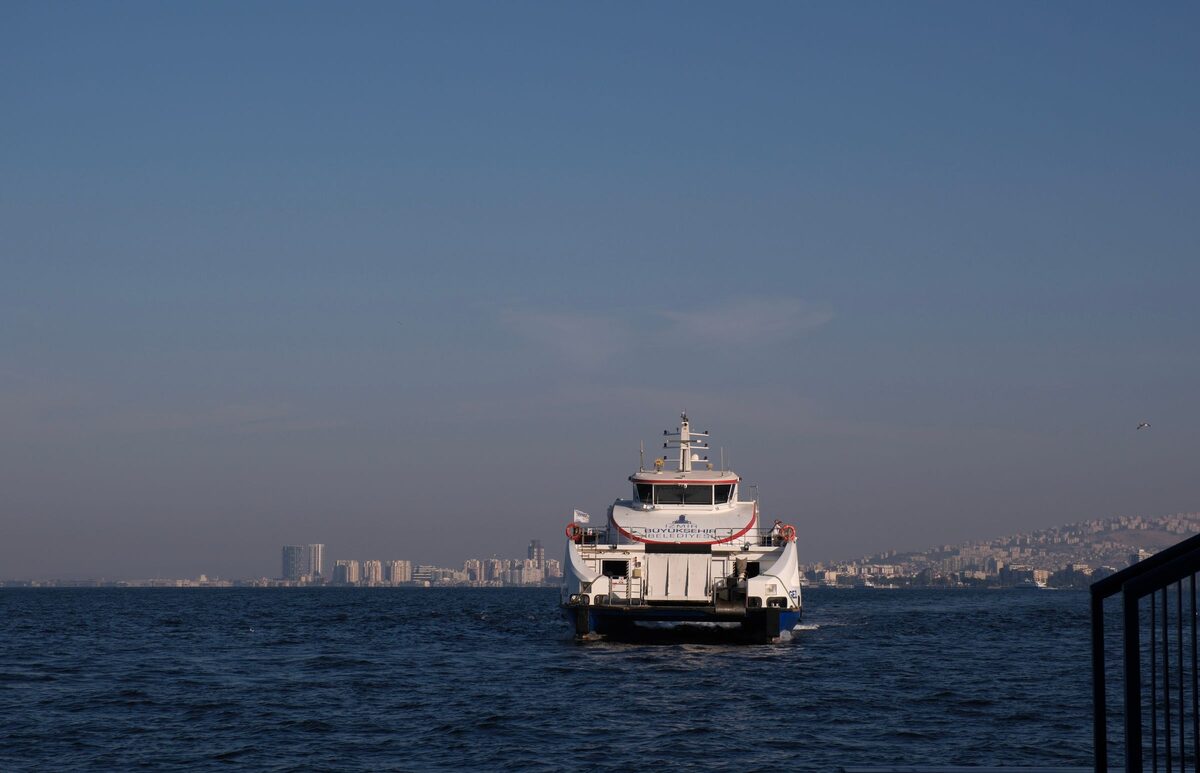 A white İzdeniz ferry crossing the gulf of İzmir towards the camera, with the city skyline behind it