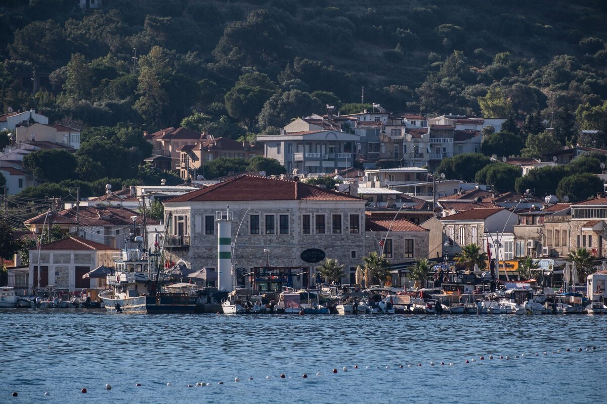 Foça's harbor with stone buildings, a small lighthouse and fishing boats along the quay, and green hills behind