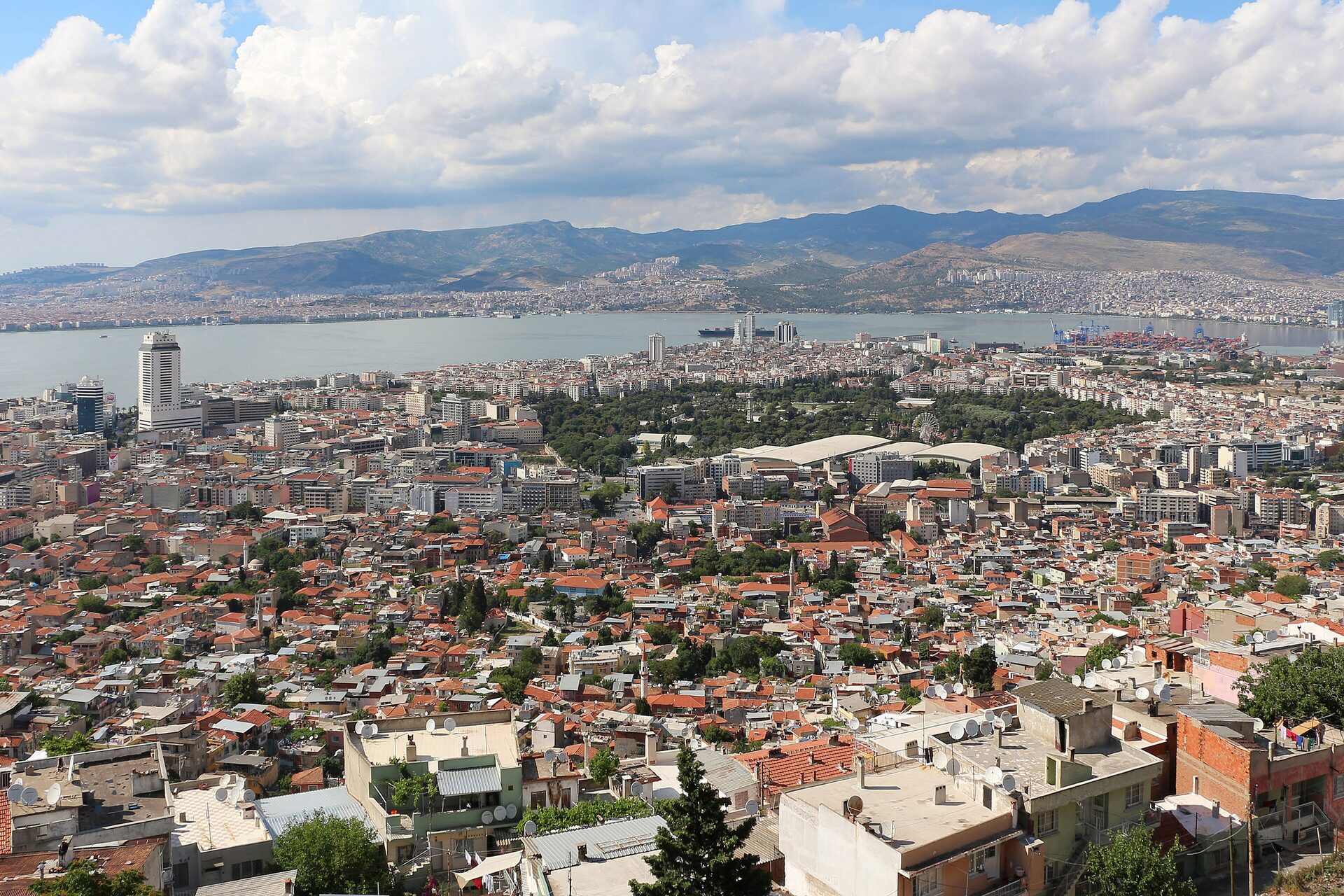 İzmir seen from Kadifekale hill: red-roofed houses running down to the green of Kültürpark, the gulf behind them and Karşıyaka and the mountains on the far shore