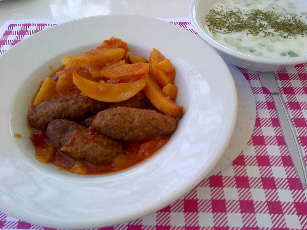 A plate of İzmir köfte, oblong meatballs baked with potato wedges and peppers in tomato sauce, with a bowl of cacık