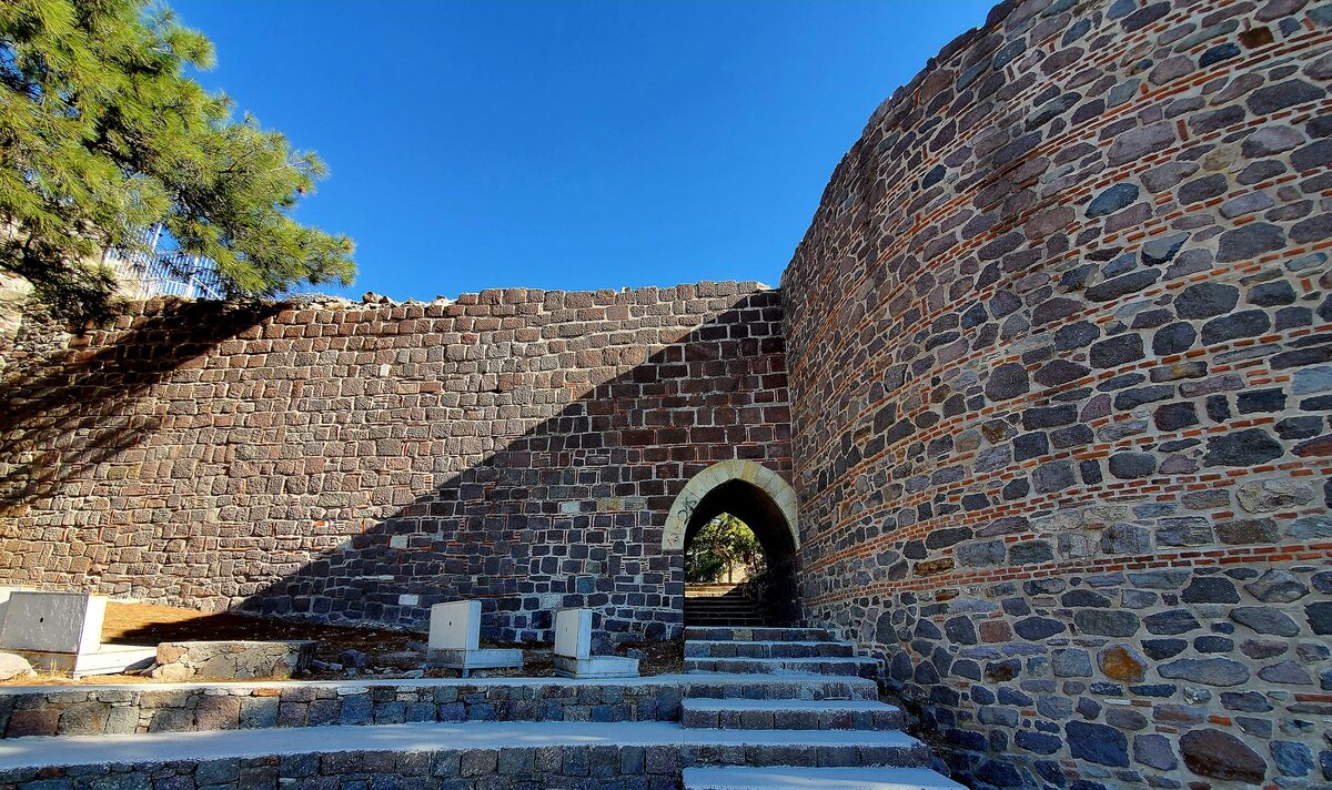The dark stone walls and a rounded tower of Kadifekale, with a small arched gate and steps under a blue sky