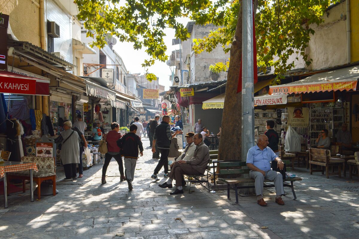A shaded lane in Kemeraltı with shop awnings, benches under a big tree and people walking past stalls